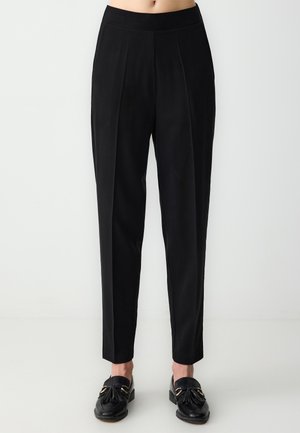 Schwarze maßgeschneiderte Hose mit einem schmalen Bund, gefaltetem Vorderteil und schmal zulaufendem Bein. Glattes Material mit matter Oberfläche, kombiniert mit schwarzen Slippern.