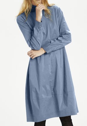 Kobieta w długiej, do kolan, stonowanego niebieskiego koloru sukience z guzikami na mankietach, pozuje z jedną ręką przy ustach, a drugą zgiętą na piersi.