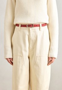 Pull côtelé crème, pantalon beige taille haute avec de grandes poches à patch, accentué par une fine ceinture en cuir rouge avec une boucle dorée circulaire.