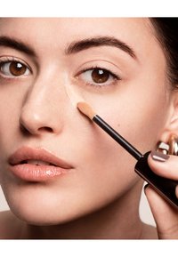 Flytende concealer i en mørk tube med applikator, påført modellens kinn, med glødende hud og en nøytral leppestiftfarge.