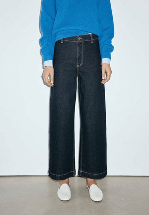 Persona con vaqueros anchos de pierna oscura, zapatos blancos sin cordones y un suéter azul, de pie contra una pared blanca.