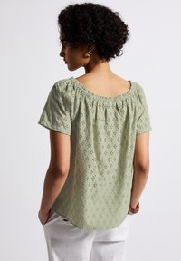 Blusa verde chiaro con maniche corte, motivo decorativo a occhiello, scollatura elastica e una vestibilità ampia, abbinata a pantaloni bianchi.