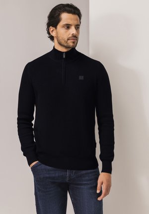 Zwarte gebreide pullover met een hoge kraag en gedeeltelijke rits, uitgerust met een gestructureerd patroon en een klein logovignet op de borst.