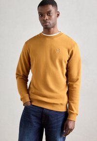 Felpa gialla senape realizzata in tessuto morbido, con scollo a girocollo e polsini a coste. Piccolo logo ricamato sul petto. Abbinata a jeans blu scuro.