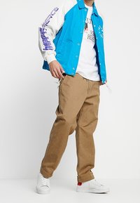 Světlomodrý větrovka s bílými rukávy, grafický text na rukávech, doplněná o béžové kalhoty a bílé tenisky s červeným akcentem.