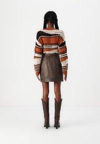 Maglione all'uncinetto con strisce arancioni, nere e crema; gonna mini in pelle marrone; stivali da cowboy marroni fino al ginocchio. Vista posteriore.
