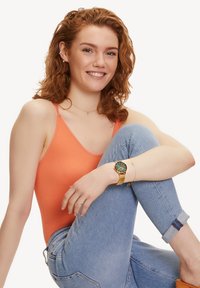Oranje geribbelde tanktop, lichtblauwe jeans en een groen horloge met gouden accenten. Glad huid, lichtbruin krullend haar, zittende houding.