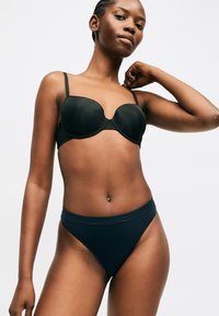 Γυναίκα που φοράει μαύρο σουτιέν και μαύρα εσώρουχα με ψηλή κοπή, πάνω σε απλό φωτεινό φόντο, ποζάροντας με ένα χέρι σηκωμένο.