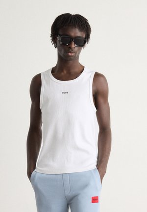 Top fără mâneci alb cu logo-ul "HUGO", asortat cu pantaloni ușor albaștri ce au o patch roșie cu "HUGO". Material neted, design casual.