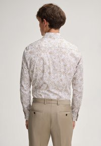 Langarmshirt mit weißem Grund, das ein verblasstes florales Muster in Beige aufweist. Der Kragen ist klassisch mit Knopfmanschetten. Kombiniert mit beigen Hosen.