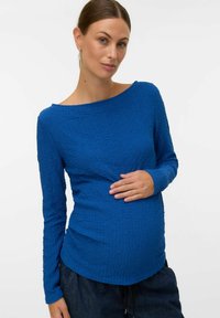 Una donna incinta in un top blu a maniche lunghe con texture accarezza delicatamente il suo pancione, in piedi contro uno sfondo bianco semplice.