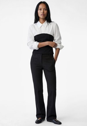 Kobieta stojąca z ręką na ramieniu, ubrana w białą koszulę na guziki pod czarnym topem bez ramiączek, czarne spodnie i czarne mokasyny.