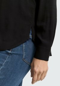 Black long-sleeve shirt with a smooth texture, side slits, worn over blue jeans. The cuff shows a subtle gold accent detail.