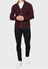 Bordový pletený kardigan s šálovým límcem a kabelovým vzorem, se třemi velkými knoflíky, v kombinaci s černými slim-fit kalhotami a bílými teniskami.