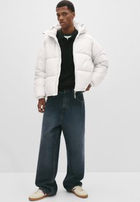 Jonge man in een witte pufferjack, zwart shirt, brede donkere jeans en beigekleurige sneakers, staande tegen een effen lichte achtergrond.
