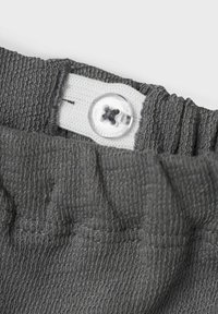 Grijze elastische tailleband met textuurstof, voorzien van een witte knoopsluiting en een zichtbare stikseldetail.