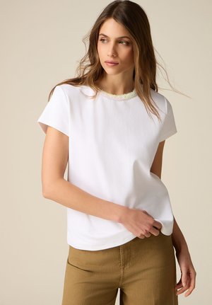 Mujer joven con largo cabello castaño, vestida con una camiseta blanca de manga corta y pantalones marrones, mirando hacia otro lado mientras ajusta el dobladillo con la mano.