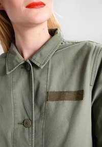Giacca verde oliva con chiusura frontale a bottoni, colletto a strati e tessuto strutturato. Presenta una toppa orizzontale in tessuto sul petto.