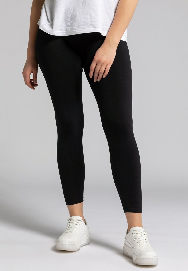 Leggings noirs en tissu extensible, avec une texture lisse et une coupe ajustée. Associés à des baskets blanches.