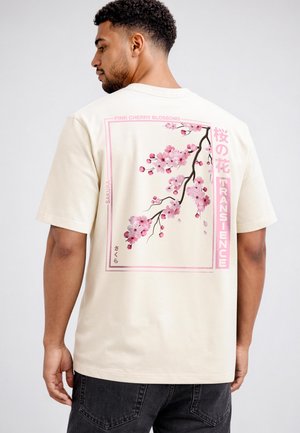 Man wearing beige t-shirt showing back with pink cherry blossom branch and Japanese text within a pink rectangular frame.