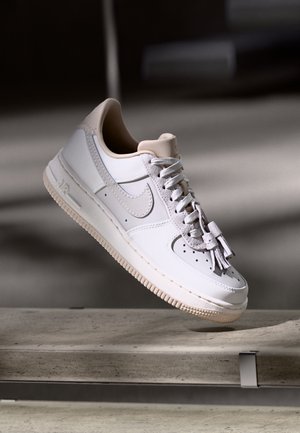 Hvid Nike Air Force 1 sneaker med lys beige sål og accenter, bundne snørebånd, svævende over en betonoverflade mod en sløret mørk baggrund.