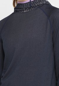 Tmavě modré sportovní triko s dlouhým rukávem, které se vyznačuje směsí materiálů, vysokým límcem a jemným vzorovaným designem podél krku a ramen.