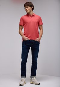 Polo shirt rose à manches courtes, associé à un jean bleu foncé. Le modèle porte des baskets gris clair avec des accents noirs. Texture de tissu lisse.