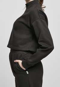 Kvinna i svart kortärmad sweatshirt och matchande byxor står sidledes med en hand i fickan mot en ljusgrå bakgrund.