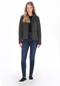 Szürke cipzáras dzseki magas gallérral, fekete inggel, sötétkék szűk farmerrel és fehér talpú fekete tornacipővel párosítva.