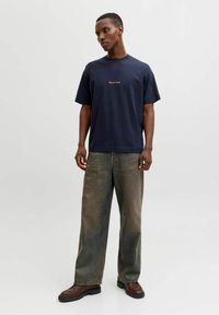 Tumesinine puuvillane T-särk lühikeste varrukatega ja ümmarguse kaelusega, millel on rinnal väike kuldne tikand; sobitamiseks on sellega laiad oliivrohelised teksapüksid.