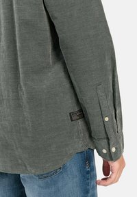 Grün karierte Hemd mit langen Ärmeln, Knopfmanschetten und abgerundetem Saum; verfügt über ein Stoffetikett an der Seite. Kombiniert mit blauen Jeans.