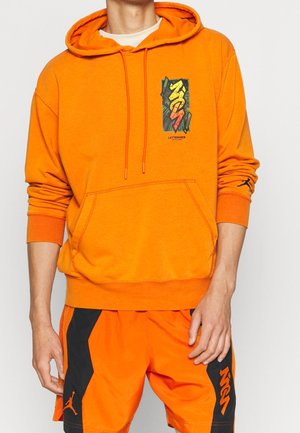 Person iført en skrigende orange hættetrøje med „Let's Dance“-grafik og matchende orange og sorte shorts med Jumpman-logo på ærmet og shortsene.