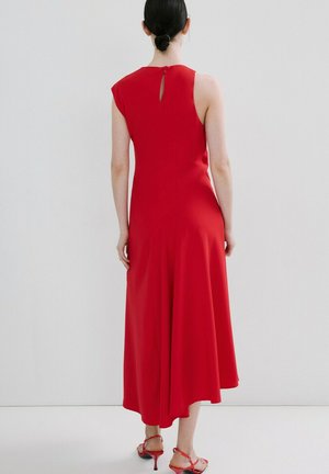 Mujer con cabello oscuro recogido en un moño, vestida con un vestido midi rojo sin mangas y sandalias de tacón rojas con tiras, de pie contra un fondo liso y claro.