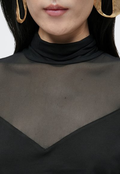 Fekete felső magas nyakú kialakítással, áttetsző hálós anyaggal a vállaknál és áttetszetlen, szilárd alapanyagból. Nagy arany fülbevalókkal kiemelve.