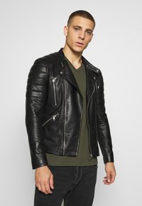 Zwarte leren motorjack met gewatteerde schouderdetails, zilveren ritsen, asymmetrische voorkant en een aansluitend ontwerp. Draag het over een groene shirt.