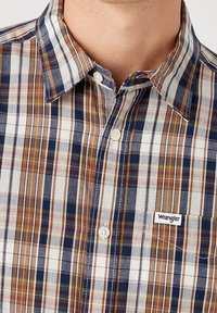 Geruite overhemd in bruin, marineblauw en wit met contrasterende texturen. Heeft een button-down kraag en het Wrangler-logo op de zak.