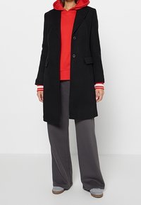 Personne portant un sweat à capuche rouge sous un long manteau noir, un pantalon gris à jambes larges et des chaussures gris clair, debout devant un fond uni.