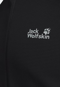 Close-up van zwart gestructureerde stof met wit Jack Wolfskin-logo en pootafdrukembleem op het oppervlak.