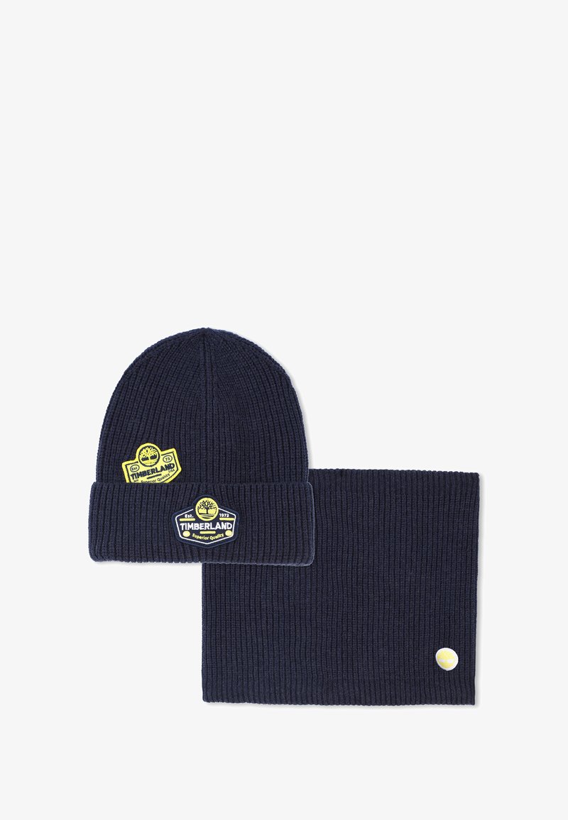 Marineblauwe ribgebreide muts en bijpassende nekwarmer, beide met gele Timberland logo patches.