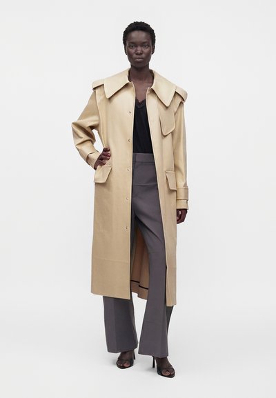 Femme portant un long manteau beige par-dessus un haut noir et un pantalon large gris, debout avec une main sur la hanche devant un fond uni.