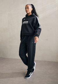 Melns kapucis ar baltu 'adidas' logo, melnas sporta bikses, baltie akcenti un melnas sporta kurpes. Gluda auduma, brīvs piegriezums, sportisks dizains.