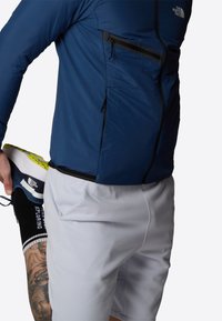 Marinblå jacka med svarta dragkedjor, grå shorts och en blå och gul sneaker, som visar upp en figursydd atletisk stil och slät textur.