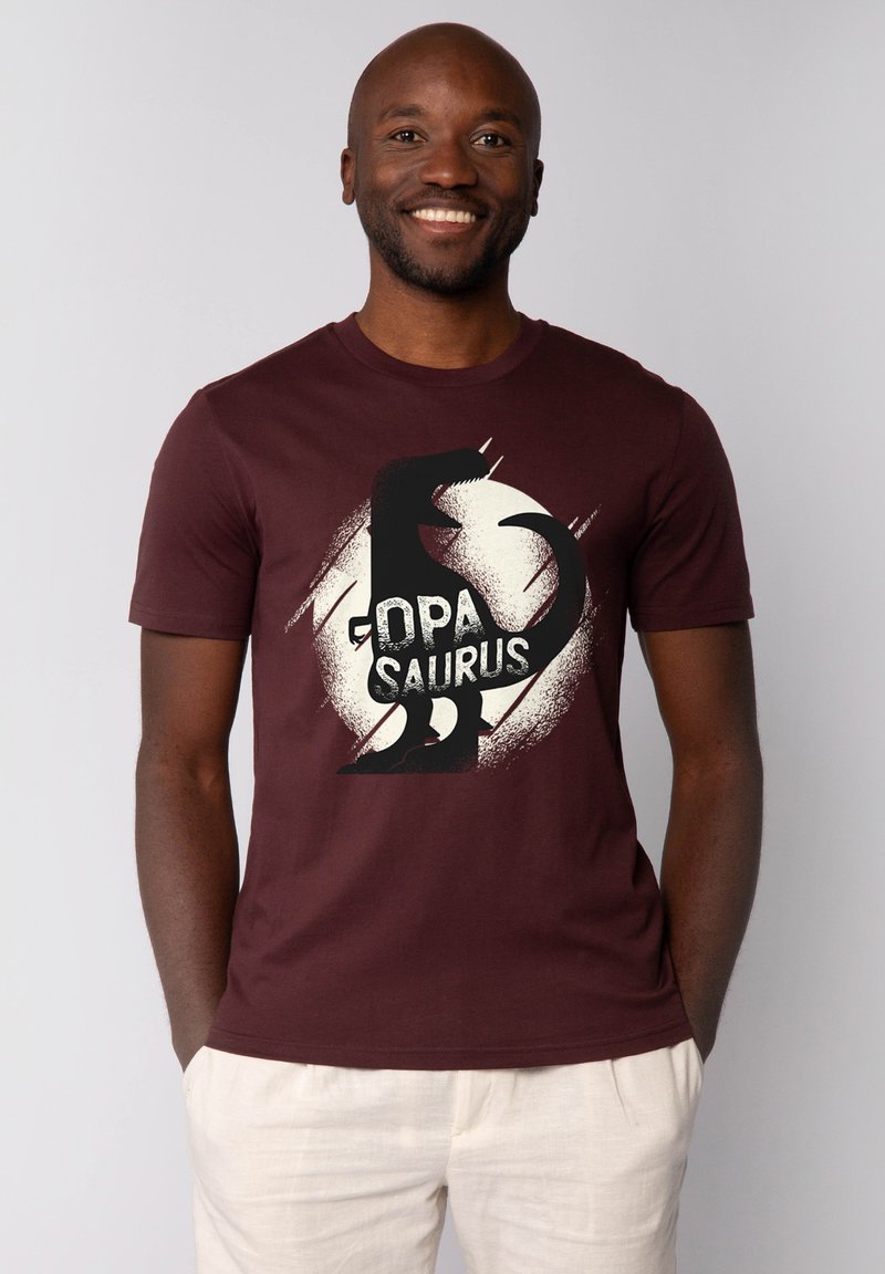 Bordeaux-rode t-shirt met een grafisch ontwerp dat een zwarte dinosaurus-silhouet toont en de tekst "OPA SAURUS," afgebeeld op een abstracte witte achtergrond.
