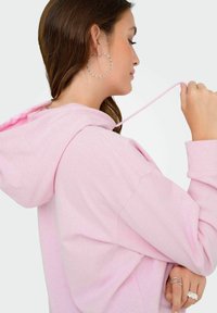 Hanorac roz deschis cu șnur, textură moale și croi casual. Asortat cu o cercei mari din argint în formă de cerc pentru un accent modern.