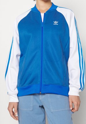 Osoba nosząca niebiesko-białą kurtkę Adidas zapinaną na zamek z paskami na rękawach oraz jasnoniebieskie spodnie, pokazana od szyi do bioder.