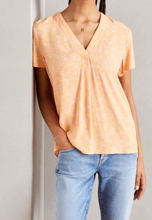 Kobieta w brzoskwiniowej bluzce z krótkim rękawem i dekoltem w szpic, z białym kwiatowym wzorem, jasnoniebieskich dżinsach, z ręką w kieszeni oraz złotym kwadratowym wisiorkiem na szyi.