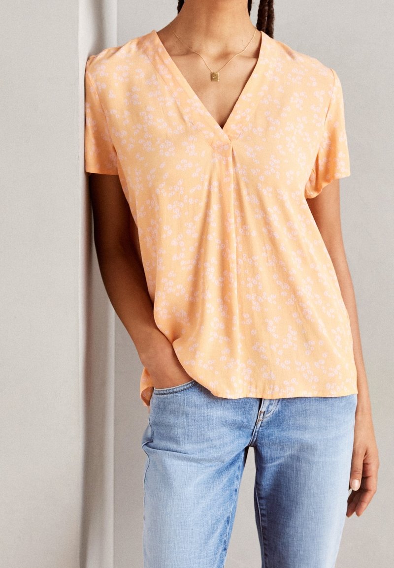 Mujer con blusa de manga corta color melocotón con estampado floral blanco y escote en V, jeans azul claro, mano en el bolsillo y collar con colgante cuadrado dorado.