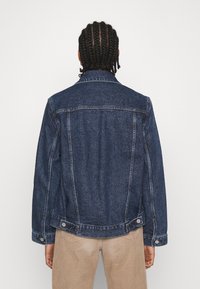 Jeansjacka i mörkblått, med en klassisk passform, knäppte manschetter och synlig sömnad. Slät textur med en något sliten yta.