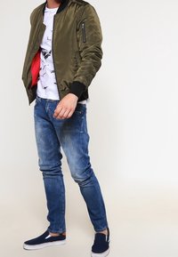 Muž v olivovém bomber jacketu přes bílém tričku s grafikou, v modrých vybledlých džínách a námořnických slip-on botách, stojící proti jednoduchému světlému pozadí.