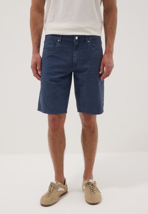 Mann trägt dunkelblaue knielange Denim-Shorts, weißes T-Shirt und beige Schnürschuhe, steht vor einem einfarbigen hellen Hintergrund.